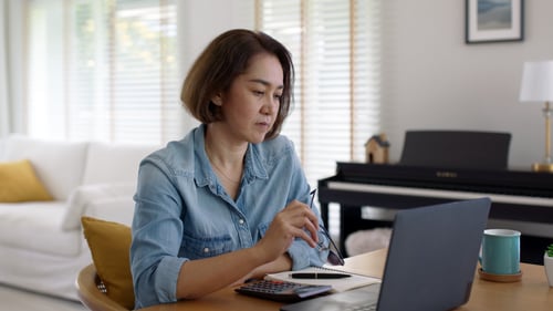 adult again woman sitting at table with calculator and notebook, staring at laptop screen