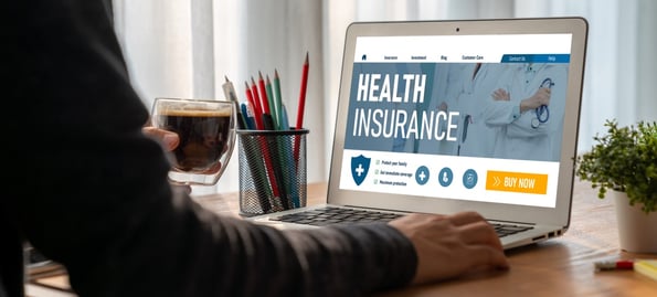 A medicare health insurance website on a laptop screen sitting on a desk while a person holding a cup of coffee reviews the information