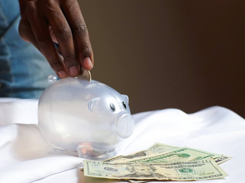 hand dropping coins into a clear piggy bank sitting on a tabletop with money