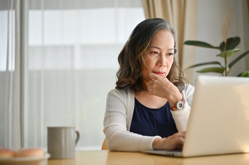 A middle aged asian woman sitting at dining room table with a cup of coffee nearby concentrating on Medicare information on laptop screen.