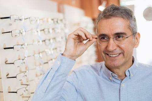 a senior man standing near rack display of many prescription eyeglass frames and smiling while trying on a pair