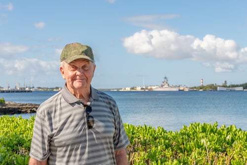 a senior man veteran in a camo hat, standing in front of water front memorial site with ships in the background
