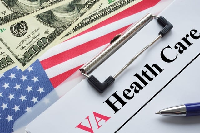 VA health care paperwork on a clipboard with American flag and hundred dollar bills in background