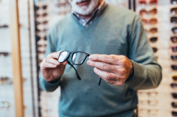 close up of senior man in sweater holding a pair of prescription eyewear with black frames inside of eye clinic with glasses and sunglasses in the background