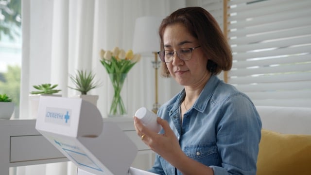 a mature asian woman sitting near table looking at a prescription medication bottle from mail order pharmacy