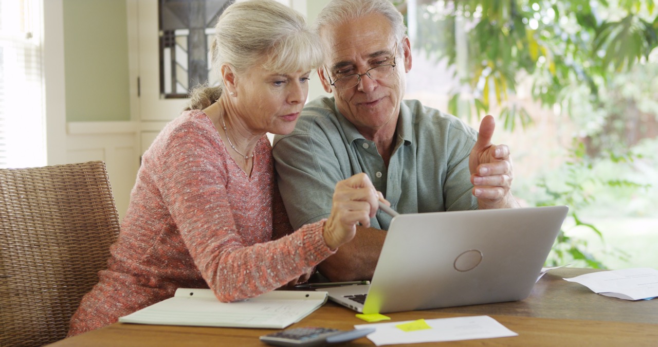 An older man and woman sit at a table reviewing their options on a laptop. They appear engaged in a discussion, with a notepad, calculator, and documents spread out in front of them, illustrating the process of researching Medicare plans.