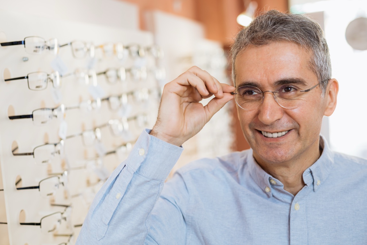 a senior man standing near rack display of many prescription eyeglass frames and smiling while trying on a pair