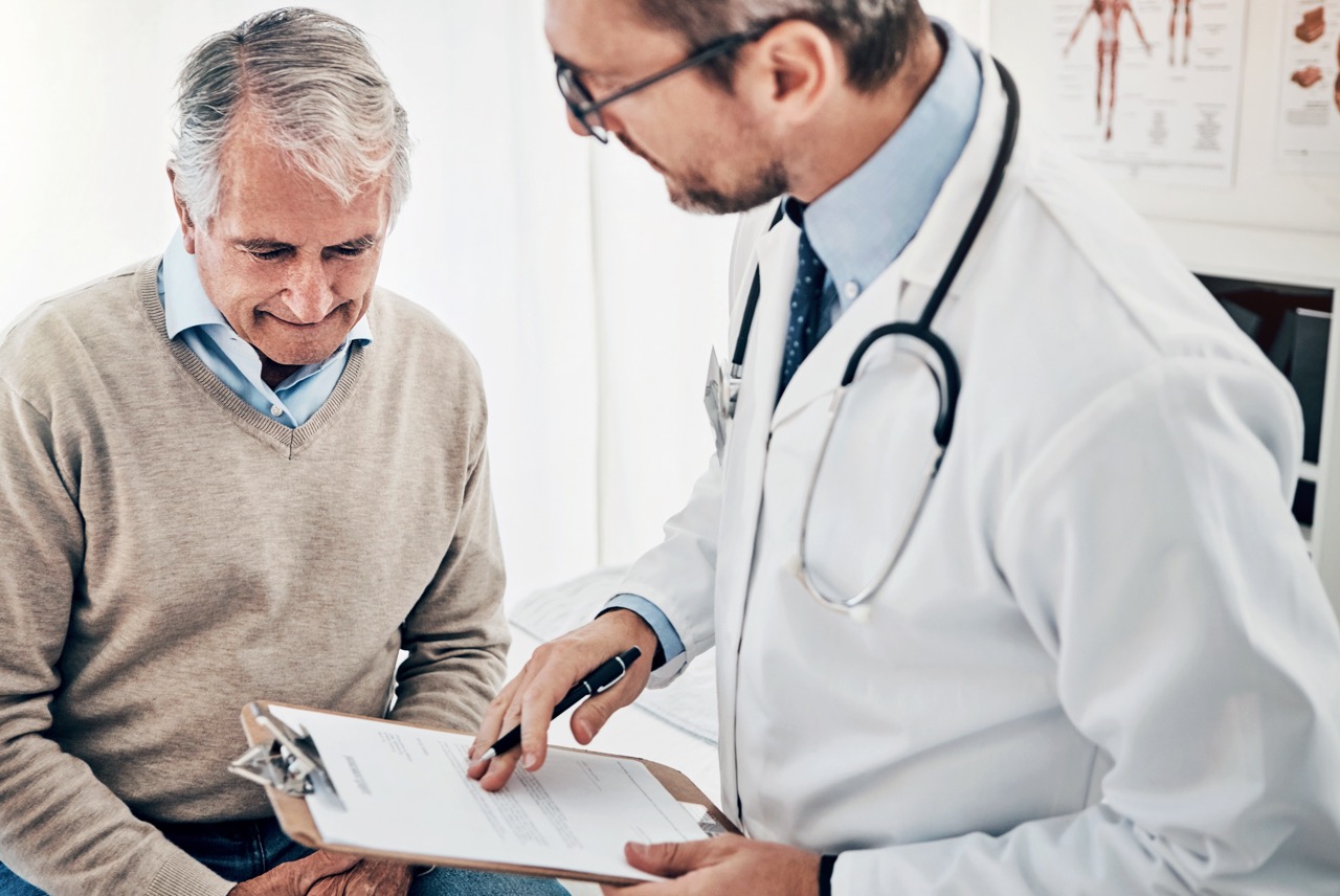 a senior man sitting in doctor's office speaking to a male medical physician holding a clipboard